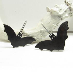 May include: A pair of black bat-shaped earrings featuring a matte finish and small silver-toned metal accents for the eyes. Each bat is suspended from a thin, dark metal ear wire hook, creating a distinct silhouette with pointed wings and ears.