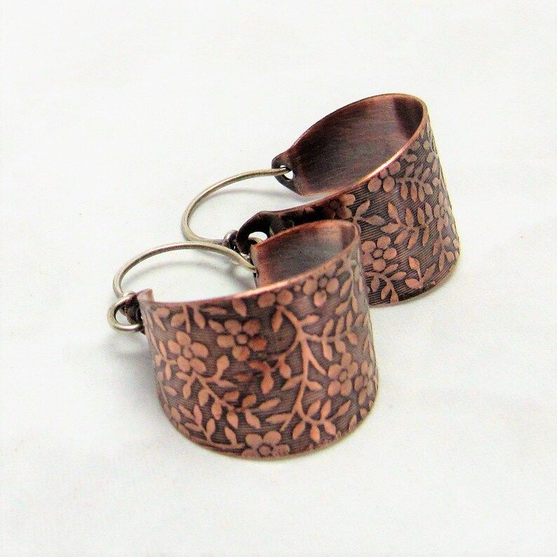 May include: A pair of copper hoop earrings with a floral design. The earrings are made of a hammered copper and have a silver hook.