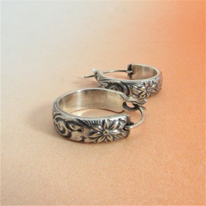 May include: A pair of silver hoop earrings with a floral design. The earrings are made of a thick, textured metal and have a simple hoop shape.