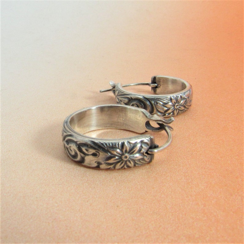 May include: A pair of silver hoop earrings with a floral design. The earrings are made of a thick, textured metal and have a simple hoop shape.