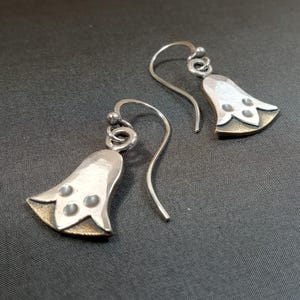 May include: A pair of silver bell-shaped earrings with a three-dot design. The earrings have a silver hook and a gold-colored base. The earrings are displayed on a dark gray surface.