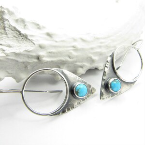 May include: A pair of silver earrings with turquoise stones. Each earring features a hammered, triangular shape with a circle cut out. The earrings have a simple hook closure.