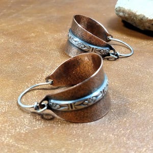 May include: A pair of copper-colored hoop earrings. Each earring features a wide, curved band with a hammered texture and a silver-colored band with a floral design. The earrings have silver-colored wire hooks.