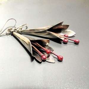 May include: A pair of silver earrings shaped like flowers with red accents. The earrings are made of metal and have a delicate, feminine design.