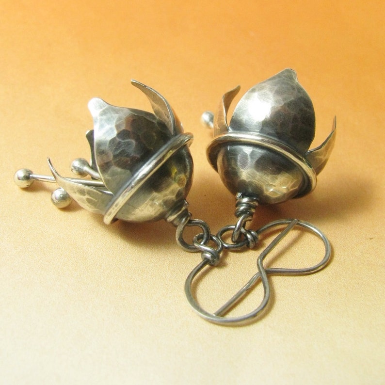 K&ouml;nnte beinhalten: Ein Paar silberne Ohrringe mit einem geh&auml;mmerten Metalldesign. Die Ohrringe sind in Form einer Bl&uuml;tenknospe mit einer kleinen Kugel am Ende jedes Stiels geformt.