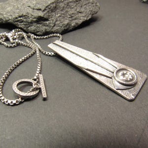 Puede incluir: Un collar de plata con un colgante geom&eacute;trico con una superficie texturizada y un dise&ntilde;o circular. El colgante tiene un dise&ntilde;o en forma de estrella en relieve en el centro.