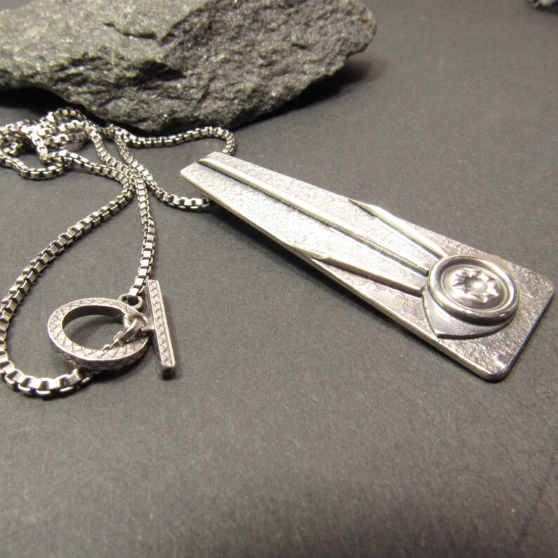 Puede incluir: Un collar de plata con un colgante geom&eacute;trico con una superficie texturizada y un dise&ntilde;o circular. El colgante tiene un dise&ntilde;o en forma de estrella en relieve en el centro.