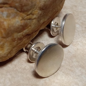 May include: Two silver-colored, round cufflinks with a polished finish. The cufflinks are displayed on a tan fabric surface, with a large, textured rock in the background. The cufflinks have a classic, minimalist design.