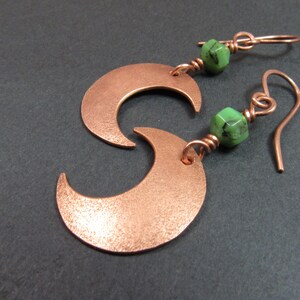 May include: Copper crescent moon earrings with green gemstone beads. The earrings feature a textured finish and are suspended from copper ear wires. The design is a celestial theme, suitable for everyday wear or special occasions.