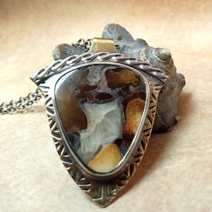 May include: A handcrafted pendant necklace featuring a triangular, patterned stone in shades of brown, tan, and white. The stone is set in a silver-toned metal frame with a decorative border and hangs from a silver chain. The pendant has a brass bail.