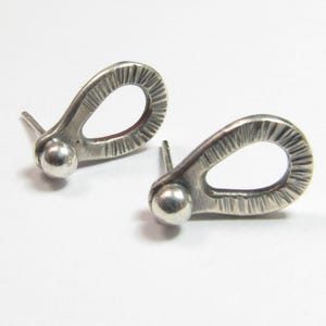 May include: A pair of silver-toned stud earrings. Each earring features a teardrop-shaped design with textured detailing and a small ball at the base. The earrings are set against a white background.