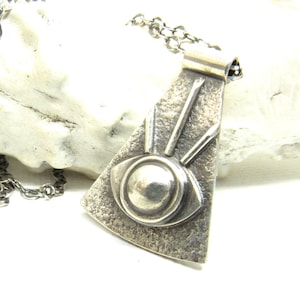 May include: A silver-toned pendant on a matching chain featuring a textured, triangular base with a central circular eye motif. Three thin, vertical silver bars extend upward from the eye design, which is set against a dark, oxidized metal surface.