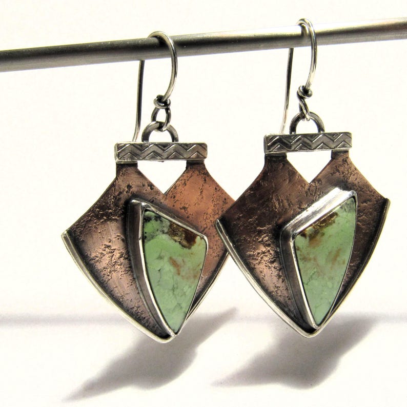 May include: A pair of silver and copper earrings with green gemstone accents. The earrings feature a geometric design with a triangular shape and a textured finish.