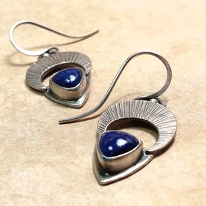 May include: A pair of silver drop earrings featuring a triangular deep blue lapis lazuli stone set in a textured metal frame. The earrings have a curved hook wire and a semi-circular top section with fine vertical etched lines.