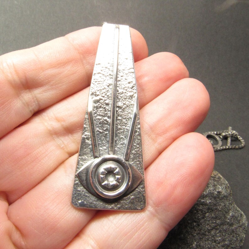 Puede incluir: Un colgante de plata con forma de tri&aacute;ngulo con un dise&ntilde;o de ojo estilizado. El colgante tiene una superficie texturizada y un ojo en relieve con un centro circular.