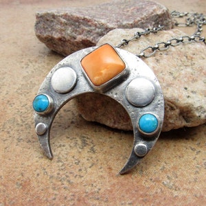 Puede incluir: Un collar con un colgante de media luna de plata, adornado con una gran piedra preciosa naranja y dos piedras turquesas más pequeñas. El colgante está decorado con pequeños círculos de plata.