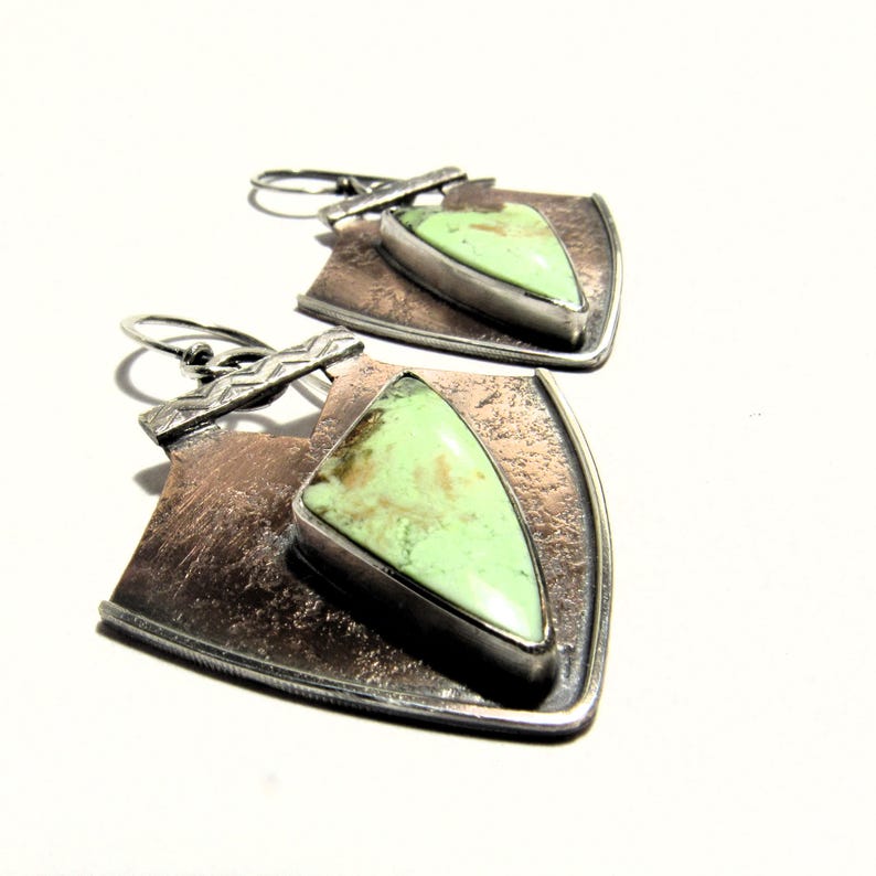 May include: A pair of silver and copper earrings with a green gemstone in the center of each earring. The earrings are shaped like a shield with a textured surface.