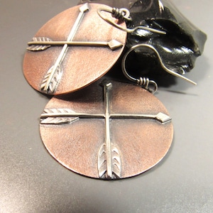 May include: A pair of copper earrings with silver arrow designs. The earrings are round and have a hammered texture.