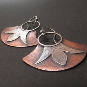 May include: A pair of dangle earrings featuring a fan-shaped copper base with a textured surface. Each earring has a silver-toned metal floral overlay attached to a circular ring, which connects to a curved ear wire for secure placement.
