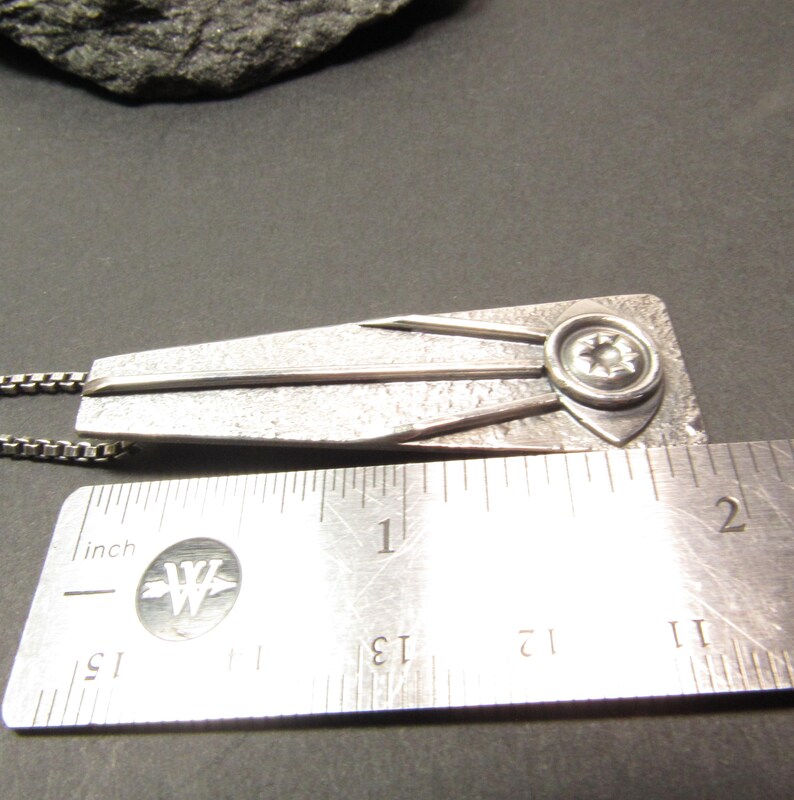 Puede incluir: Un collar con un colgante de plata con un dise&ntilde;o geom&eacute;trico y una estrella en el centro. El colgante mide aproximadamente 3,8 cm de largo.