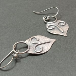 May include: A pair of silver-toned drop earrings featuring a textured, leaf-shaped pendant with a raised, swirling wire design. Each earring is attached to a simple circular ring and a curved ear wire with a small ball detail at the end.