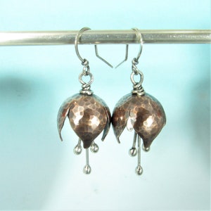 May include: A pair of hammered copper earrings with silver accents. The earrings are shaped like bells with small silver balls hanging from the bottom.