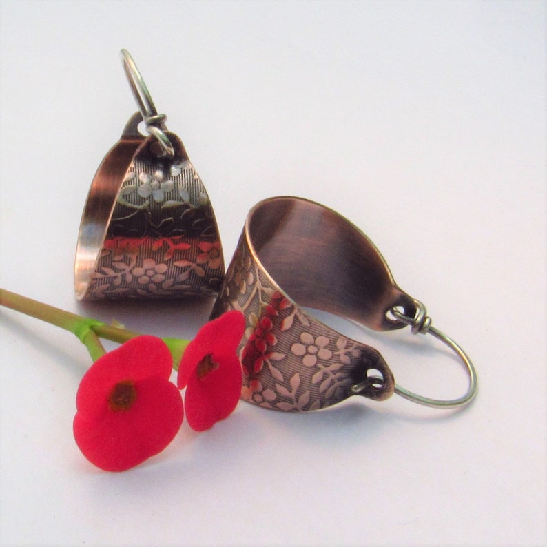 May include: A pair of copper hoop earrings with a floral pattern. The earrings are a light copper color and have a hammered finish. The floral pattern is embossed on the front of the earrings.