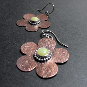 May include: A pair of flower-shaped earrings. The petals are a textured copper color, and each earring features a light green stone set in a silver-colored beaded frame. The ear wires are silver-colored.