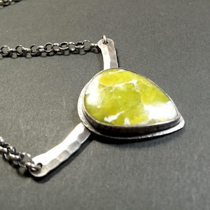 May include: A silver necklace featuring a pear-shaped, lime green stone with white marbling. The stone is set in a silver bezel and connected to a hammered silver bar and a chain. The necklace is displayed against a dark background.
