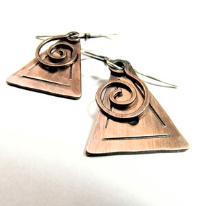 May include: Copper-colored earrings with a triangular base and a spiral design. The earrings have a geometric shape with etched lines and silver-colored ear wires. The jewelry is displayed against a white background.