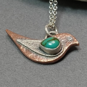 May include: A bird-shaped pendant necklace featuring a textured copper body and a silver wing overlay. A teardrop-shaped turquoise stone with brown veining is set in the center of the bird. The pendant hangs from a delicate silver chain link.