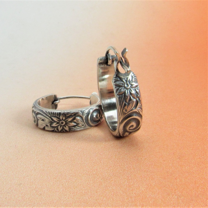 May include: A pair of silver hoop earrings with a floral design. The earrings are made of a smooth, shiny metal and have a delicate, intricate design.