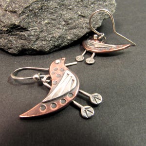 May include: A pair of copper and silver bird earrings with a geometric design. The birds have a silver wing and a copper body with silver dots. The earrings hang from silver hooks.
