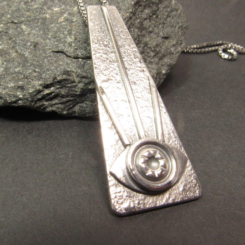 Puede incluir: Un collar con un colgante de plata con un dise&ntilde;o geom&eacute;trico texturizado. El colgante presenta un ojo estilizado con una estrella en el centro.