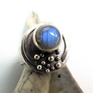 Puede incluir: Un anillo de plata con una piedra preciosa de labradorita azul engastada en una banda de plata texturizada. La banda está decorada con pequeñas cuentas de plata.