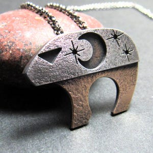May include: A silver and copper-toned pendant necklace featuring a stylized bear design. The bear's head has a crescent moon and star cutouts. The pendant is suspended from a delicate silver chain. The necklace is displayed on a reddish-brown stone.