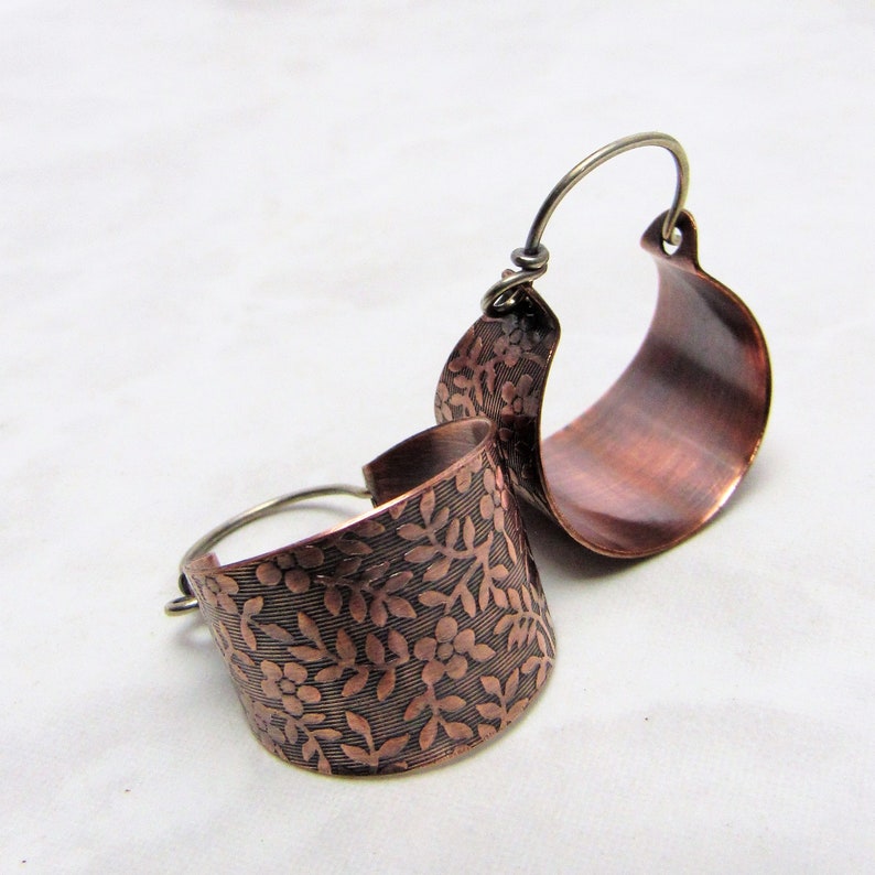 May include: A pair of copper hoop earrings with a floral pattern. The earrings are made of a hammered copper and have a slightly curved shape.