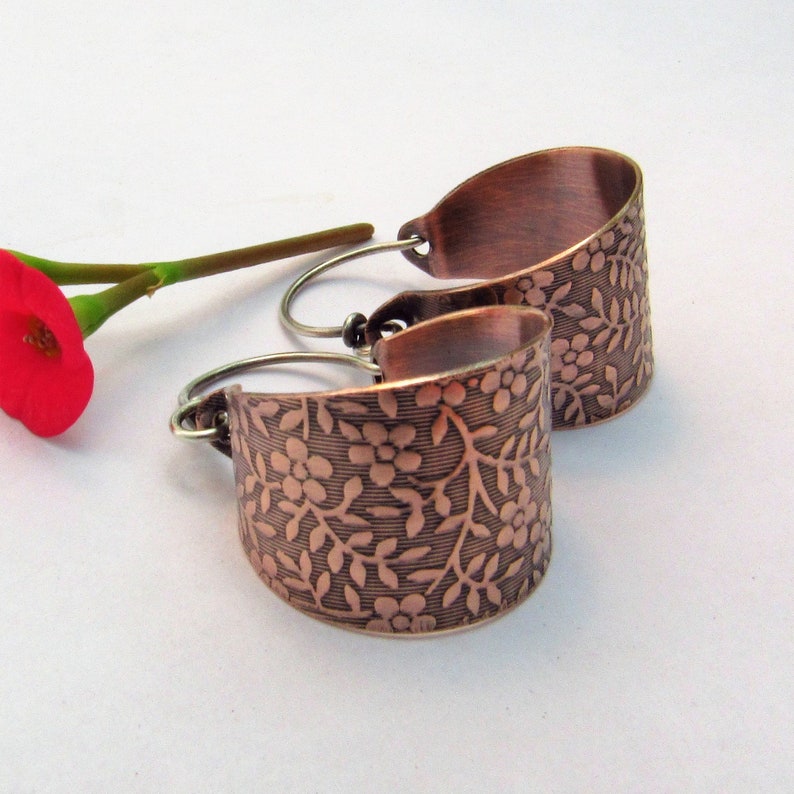 May include: A pair of copper hoop earrings with a floral design. The earrings are a light copper color and have a hammered finish.