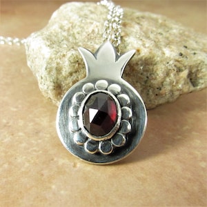 Puede incluir: Un collar con un colgante de plata en forma de granada adornado con una piedra preciosa roja. El colgante está detallado con una superficie texturizada y una parte superior en forma de corona.