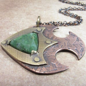 May include: A handcrafted pendant necklace featuring a large, faceted green gemstone set in a geometric brass and copper design. The pendant has a textured surface and is suspended from a dark chain. The overall style is rustic and artistic.