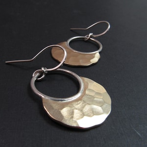May include: A pair of handcrafted earrings with a hammered gold-tone disc and a silver-tone hoop. The earrings have a textured surface and a hook closure. The jewelry is displayed on a dark background.