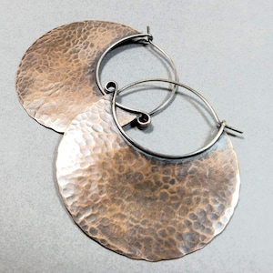 May include: A pair of hammered copper hoop earrings. The earrings feature a large, textured, circular copper pendant attached to a silver-colored hoop. The copper has a warm, reddish-brown hue, and the hoops have a simple, elegant design.