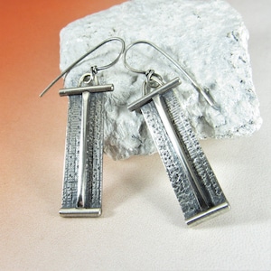 May include: A pair of silver dangle earrings with a textured rectangular design. The earrings have a simple, minimalist style.