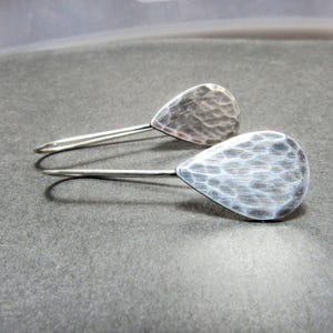 May include: A pair of silver-toned drop earrings featuring a teardrop-shaped design with a hammered, textured surface. Each earring is attached to a curved wire hook, showcasing a metallic finish with subtle light and dark shading across the metal.