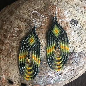 May include: A pair of dangle earrings with a black, green, and yellow beaded design. The earrings feature a teardrop shape with a crystal bead in the center.