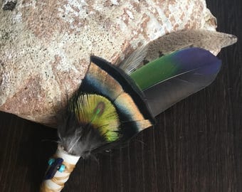 May include: A small feather smudge stick with a variety of feathers, including a peacock feather with green, blue, and yellow colours. The stick is wrapped in brown leather and has turquoise beads.