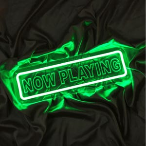 May include: A neon sign with the words "NOW PLAYING" in bright green. The sign is rectangular with a white border and is set against a black, draped fabric background. The neon glow creates a vibrant and eye-catching display.