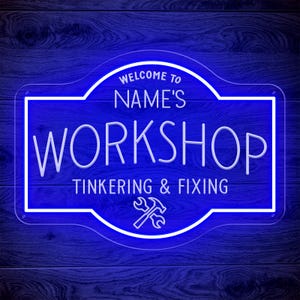 May include: A blue neon sign with white lettering that reads "WELCOME TO NAME'S WORKSHOP TINKERING & FIXING". The sign features a hammer and wrench crossed at the bottom. The sign is mounted on a dark blue wooden background.