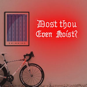 Puede incluir: Un letrero de ne&oacute;n rojo con el texto "Dost thou Even Hoist?" en fuente g&oacute;tica. A la izquierda, una impresi&oacute;n enmarcada con la palabra "SWIMMING". Una bicicleta de carretera blanca y roja en primer plano.
