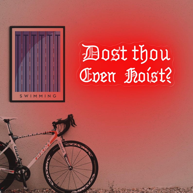 Puede incluir: Un letrero de ne&oacute;n rojo con el texto "Dost thou Even Hoist?" en fuente g&oacute;tica. A la izquierda, una impresi&oacute;n enmarcada con la palabra "SWIMMING". Una bicicleta de carretera blanca y roja en primer plano.